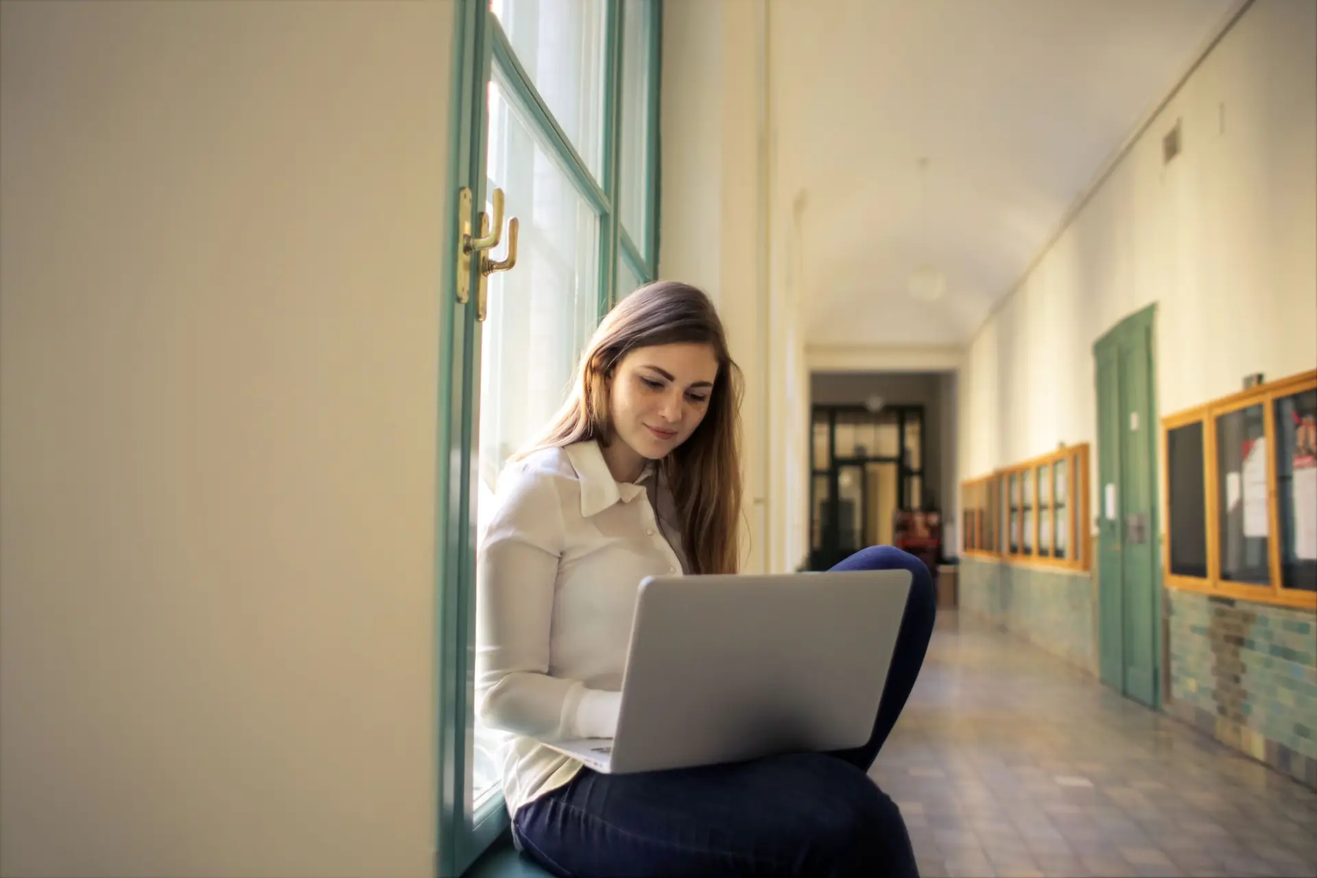 Joven sentada junto a una ventana en un pasillo antiguo trabaja con una laptop sobre las piernas