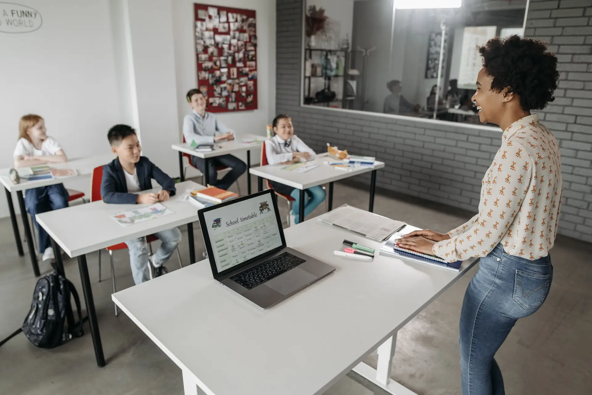 Una docente sonríe frente a un grupo de alumnos sentados en pupitres de un aula, con un horario escolar en una computadora portátil