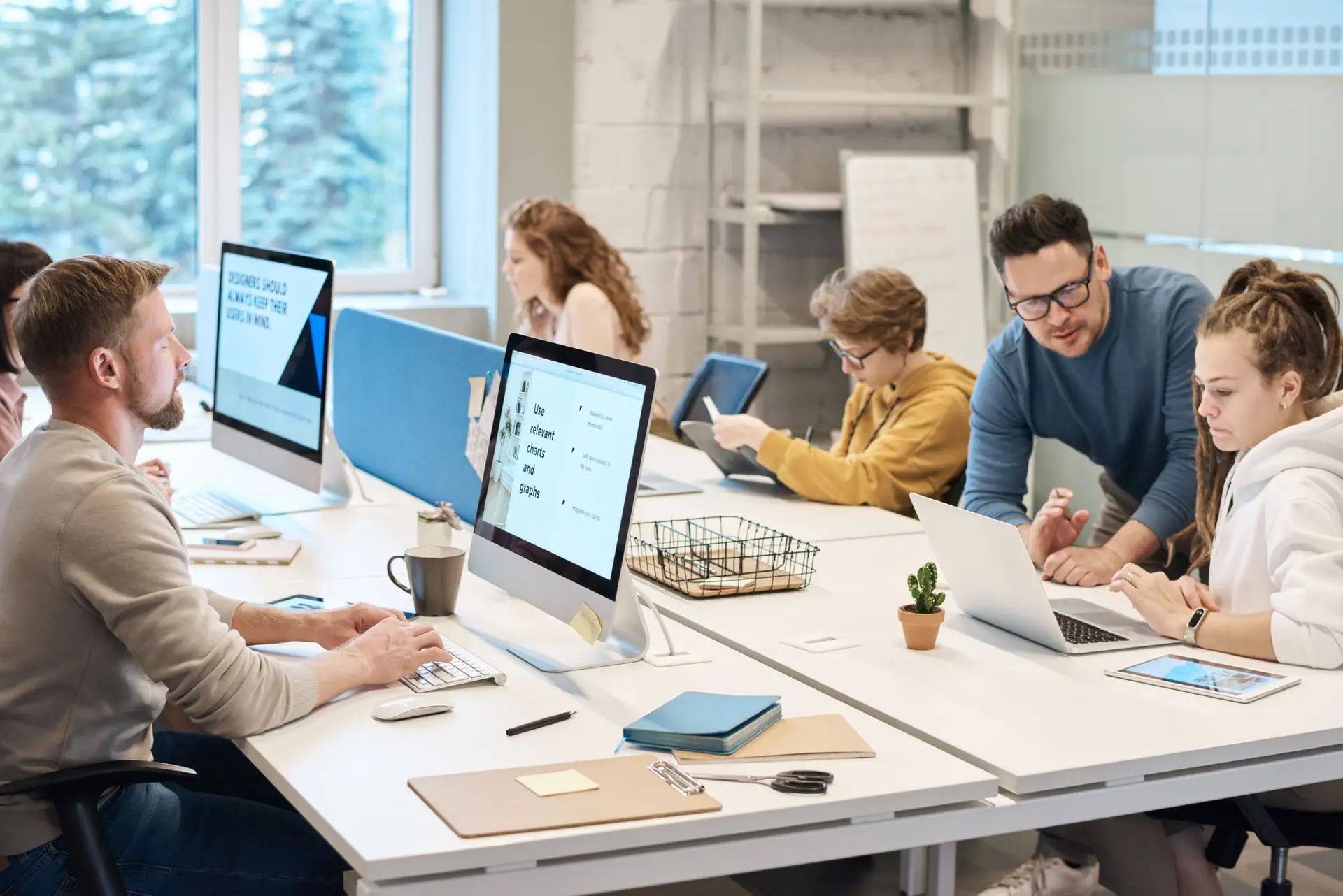 Equipo de personas trabajando con computadores en una oficina luminosa mientras un hombre ayuda a una joven con su portátil