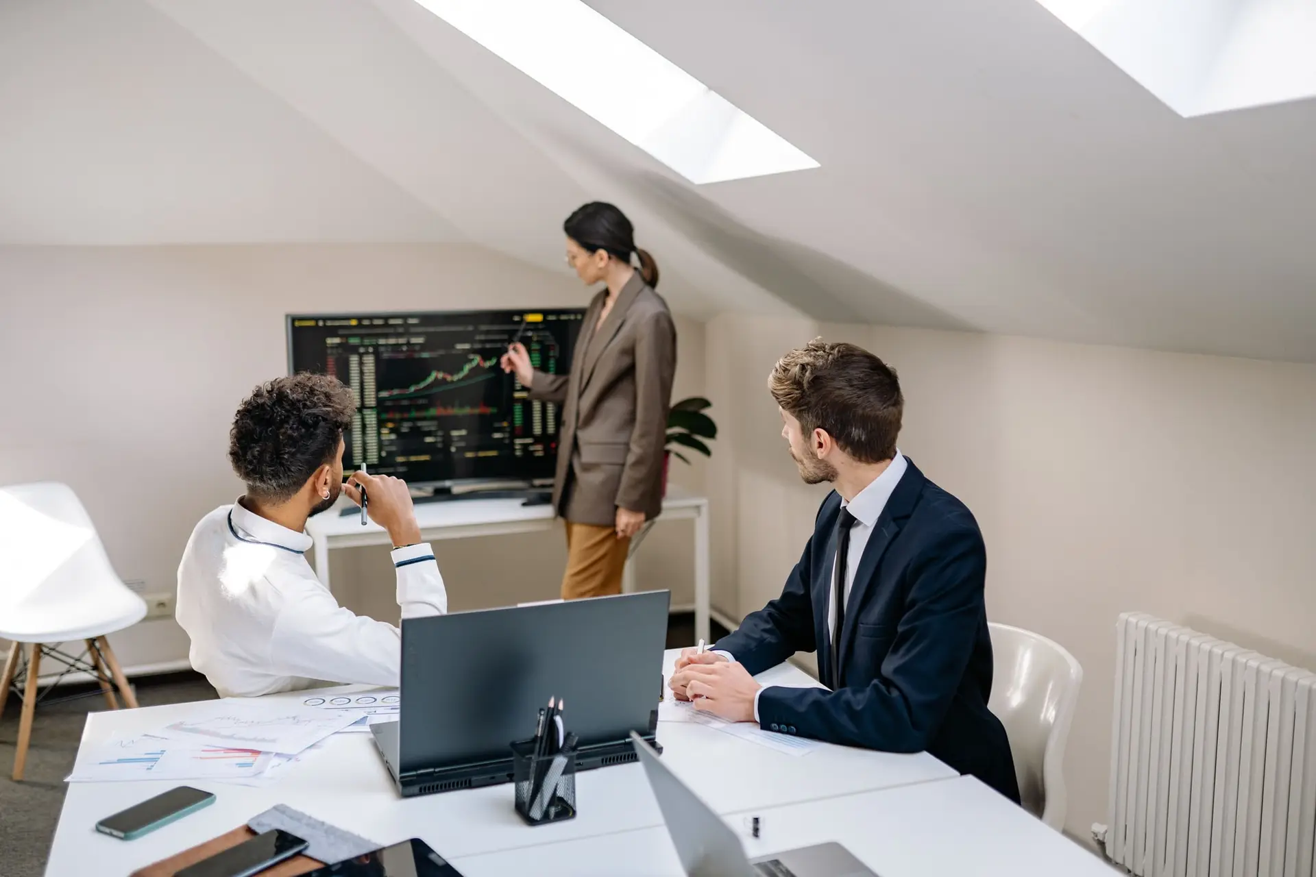 Tres profesionales en una oficina luminosa observan gráficos de datos en una pantalla grande mientras una mujer los señala