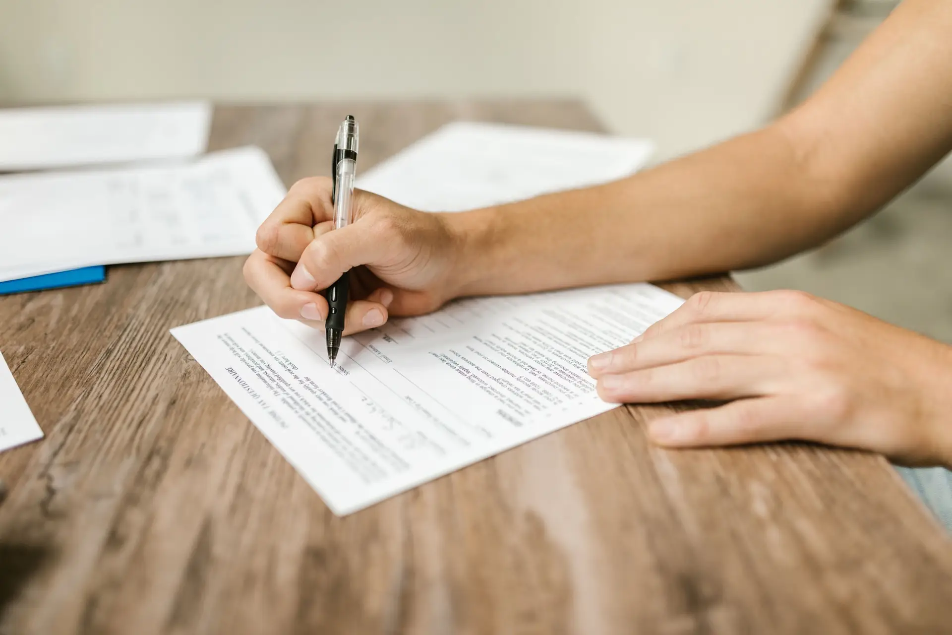 Manos de una persona llenando con un bolígrafo un formulario impreso sobre un escritorio blanco junto a otros documentos